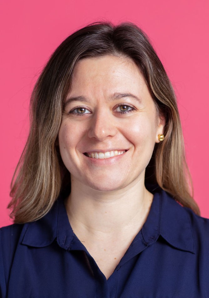 A woman in a blue shirt smiling in front of a pink background.